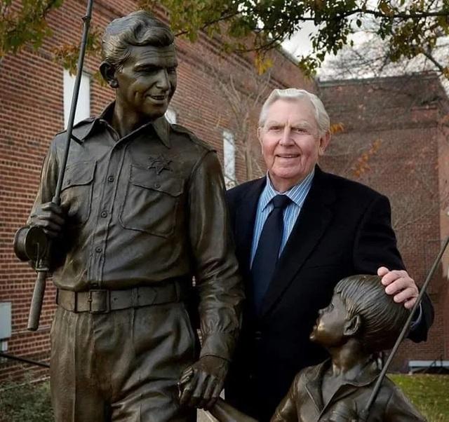Andy Griffith standing proudly next to the bronze statue of Andy Taylor and Opie in Mount Airy, North Carolina, a tribute to the beloved characters from The Andy Griffith Show.